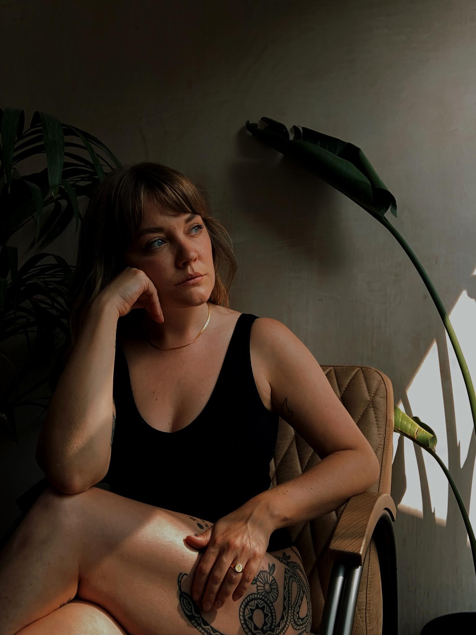 Lyzi sitting on a vintage chair in front of a plaster wall and large plants. Wearing simple black underwear and a Daisy x Estee Lalonde snake chain and forget me not signet ring.
